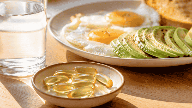 Fish oil softgel capsules beside a breakfast plate with eggs and avocado representing the recommendation to take omega-3 fish oil with a fat-containing meal for best absorption